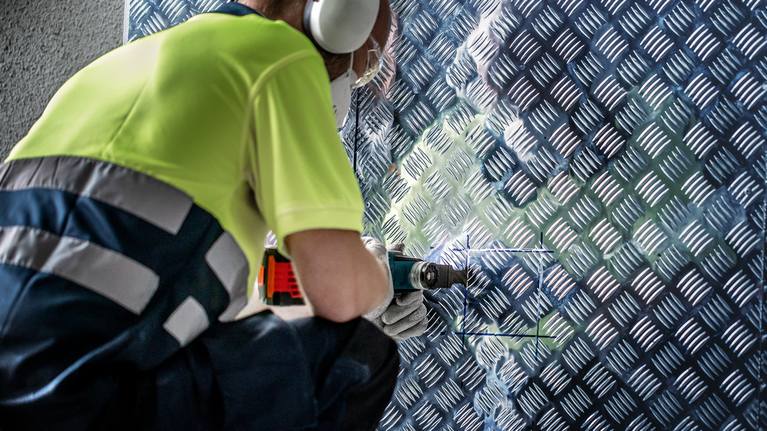 EXPERT MultiMax MAII 32 APIT Multitool Blade 32 mm Person wearing safety equipment drills into a metal patterned wall panel.