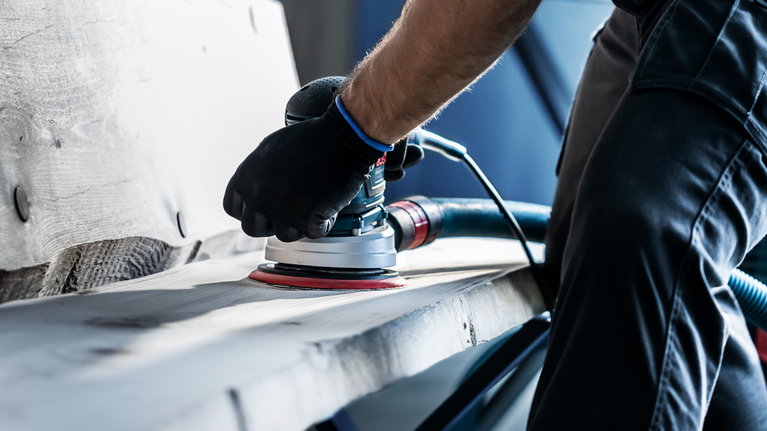 EXPERT M480 Sanding Net for Random Orbital Sanders 125 mm, G 80 5-pc Person wearing safety equipment sands a wooden plank with a power sander.