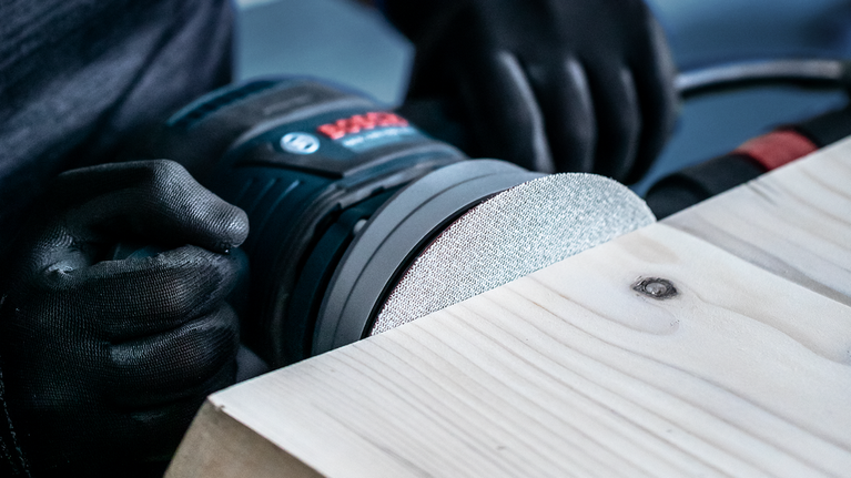 EXPERT M480 Sanding Net for Random Orbital Sanders 125 mm, G 80 5-pc Person wearing safety equipment sands a wooden surface with a power tool.