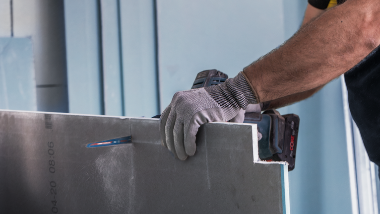 Person wearing safety equipment cuts drywall with a power tool.