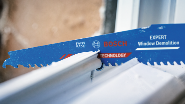 Reciprocating saw cuts through metal window frame beside a rough wall.