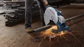 A person wearing safety equipment cuts metal bars with a chop saw, producing sparks.