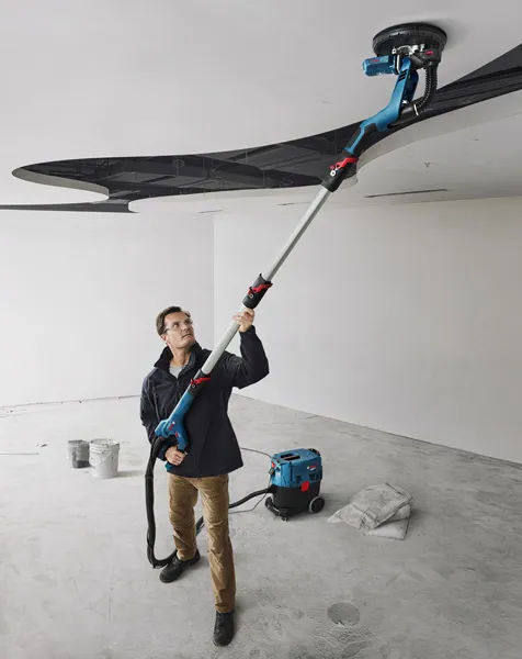 GTR 55-225 A person wearing safety equipment sands a ceiling with a drywall sander in a renovation space.