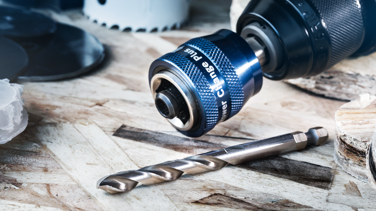 Close-up of a drill chuck attachment with a metal drill bit on a wooden surface.