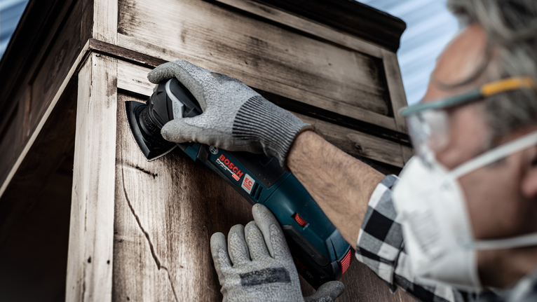 Person wearing safety equipment sands a wooden cabinet using a power tool.