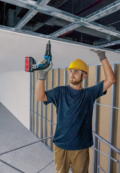 A person wearing safety equipment installs ceiling panels using a power screwdriver.