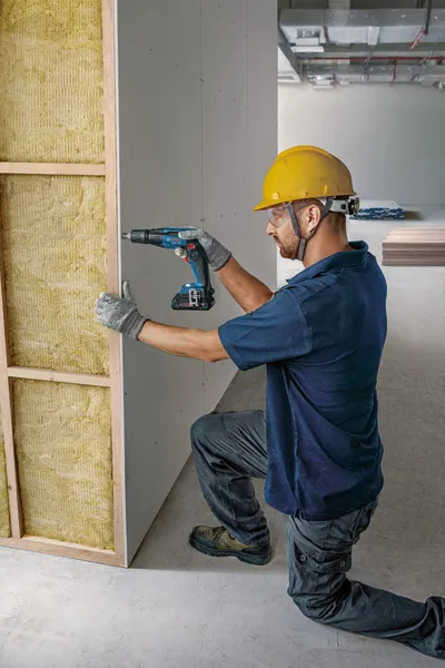 Person wearing safety equipment fastens drywall to a frame using a cordless drill.