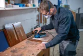 A person wearing safety equipment sands a wooden board with a random orbit sander.