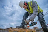 A person wearing safety equipment uses a cordless angle grinder to cut metal rebar.