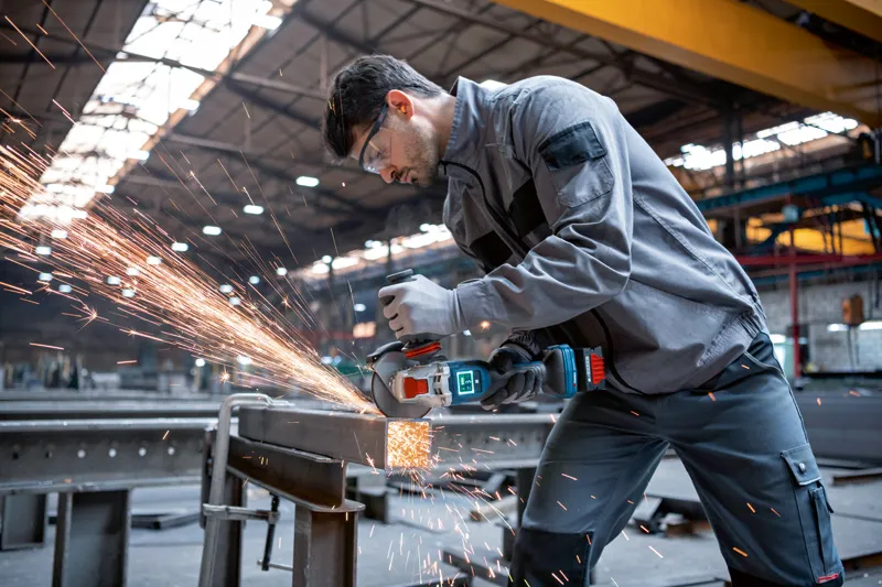 A person wearing safety equipment grinds metal beams with a cordless angle grinder.