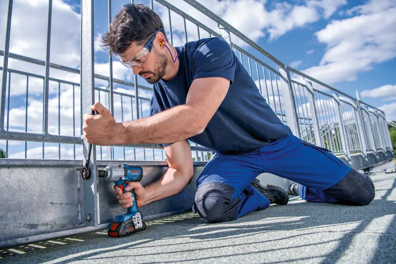 A person wearing safety equipment fastens a metal fence with a cordless impact wrench.