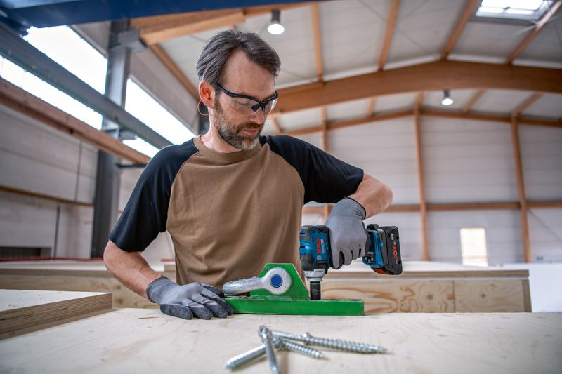 A person wearing safety equipment uses a cordless drill on a wooden workbench.