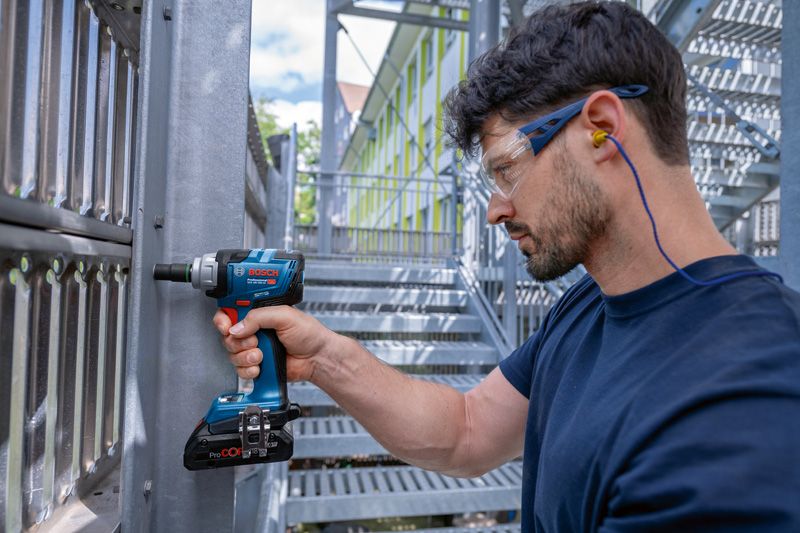 A person wearing safety equipment uses a cordless impact wrench on a metal structure.