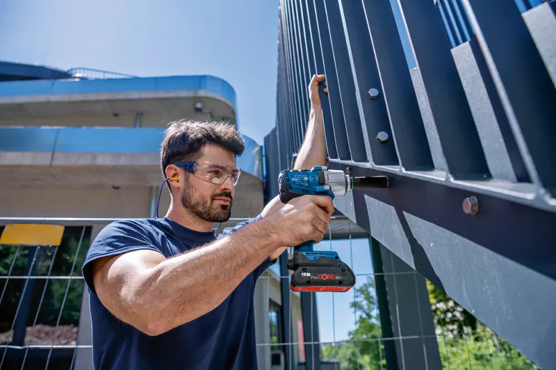 A person wearing safety equipment uses a cordless impact wrench on a metal fence.