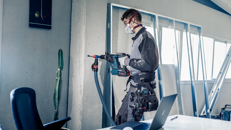 EXPERT SDS Clean max Connector for Hammer Drill Bit Person wearing safety equipment drills into a concrete wall in an indoor workspace.