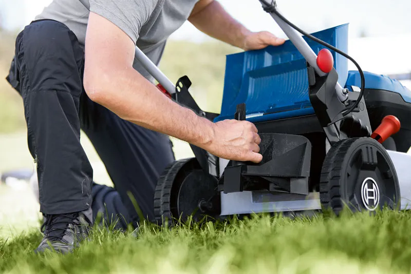 A person adjusts the compartment of a cordless lawnmower on a grassy lawn.