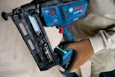 Person wearing safety equipment holds a cordless wood nailer on a light wood floor.