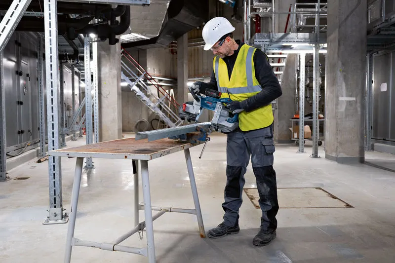 A person wearing safety equipment cuts a metal beam with a cordless band saw.