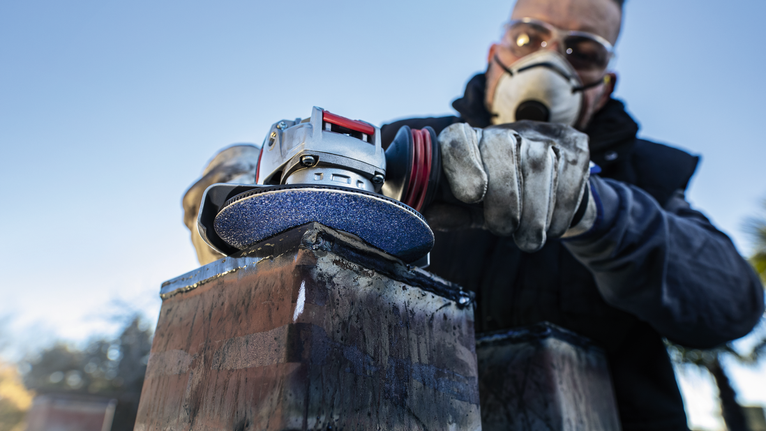 A person wearing safety equipment grinds a metal beam with an angle grinder.