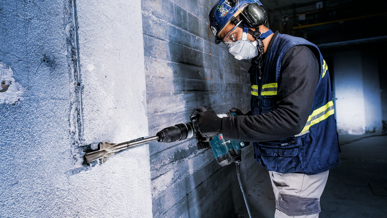 A person wearing safety equipment uses a power drill to chisel concrete.