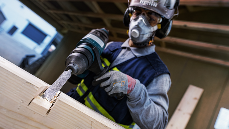Person wearing safety equipment chisels a wooden beam with a power tool.