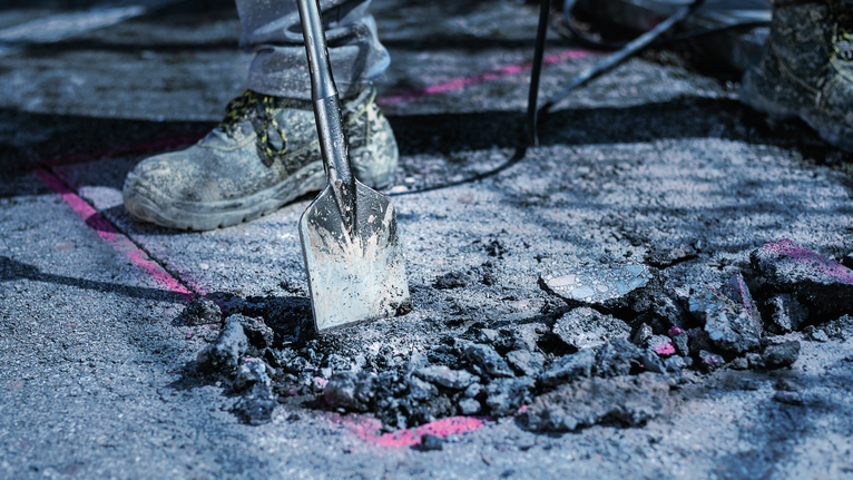 Person wearing safety equipment breaks asphalt with a demolition tool.