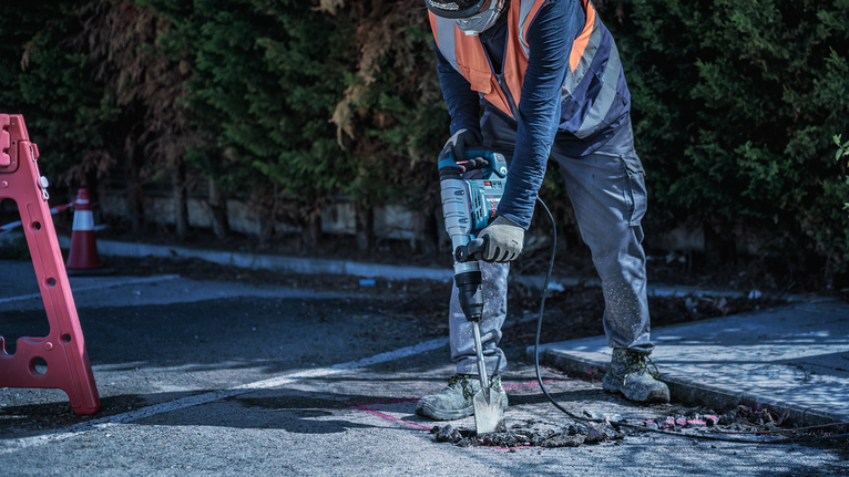 A person wearing safety equipment breaks asphalt with a power demolition tool.