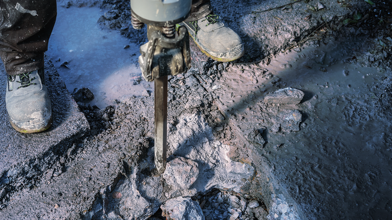 Person wearing safety equipment breaking concrete with a demolition hammer.