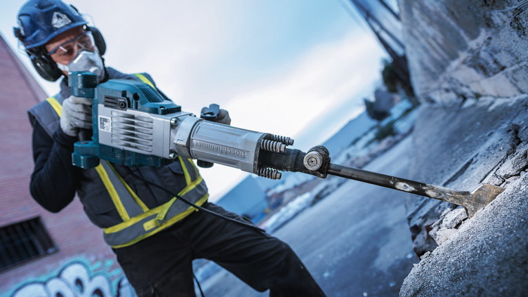 A person wearing safety equipment uses a demolition tool to break concrete.