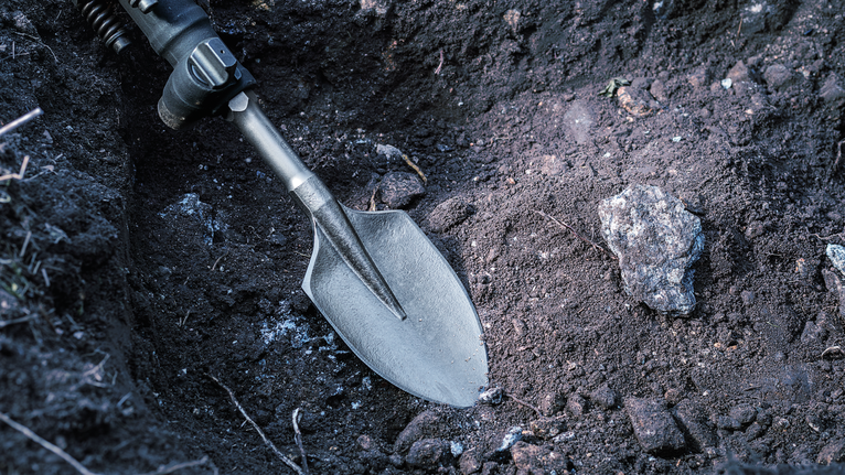 A power tool with a spade attachment digs soil near a rock.