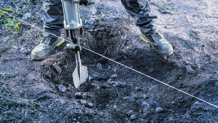 A person wearing safety equipment uses a demolition hammer to break up soil.