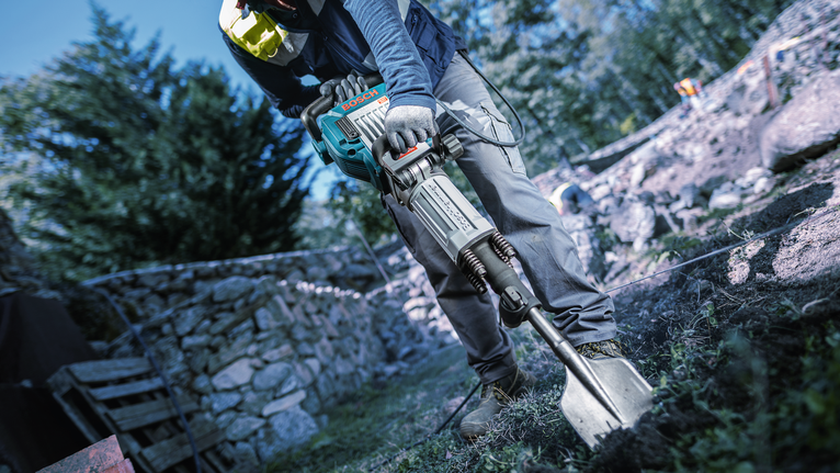 A person wearing safety equipment uses a demolition hammer to break ground outdoors.