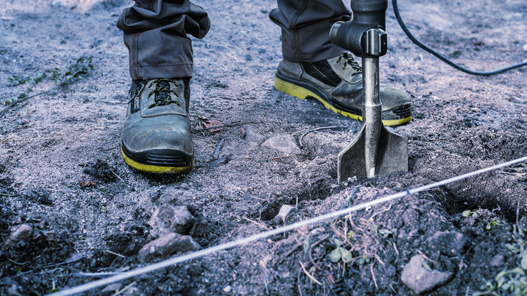 A person wearing safety equipment drills into soil with a power auger.