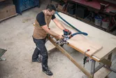 A person wearing safety equipment uses a cordless palm router on a wooden board.