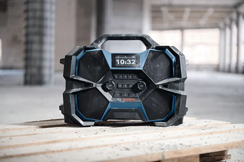 Portable construction site radio with digital display sits on wooden planks in a building under construction.
