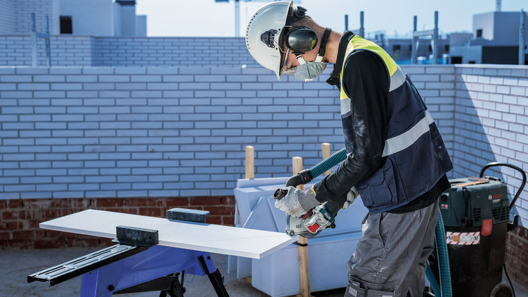A person wearing safety equipment uses a power tool to cut a board outdoors.