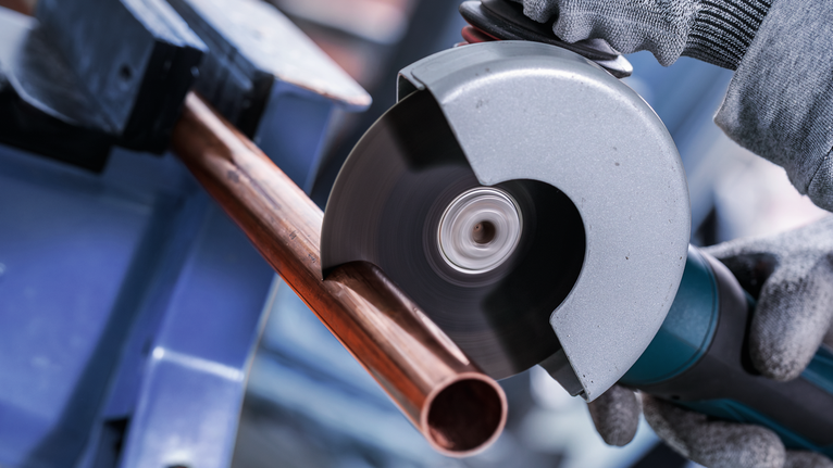 Person wearing safety equipment cuts a copper pipe with an angle grinder.
