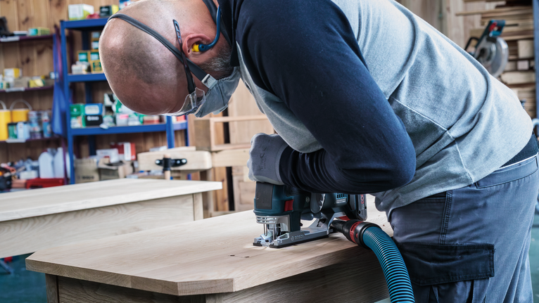A person wearing safety equipment uses a jigsaw to cut wood on a workbench.