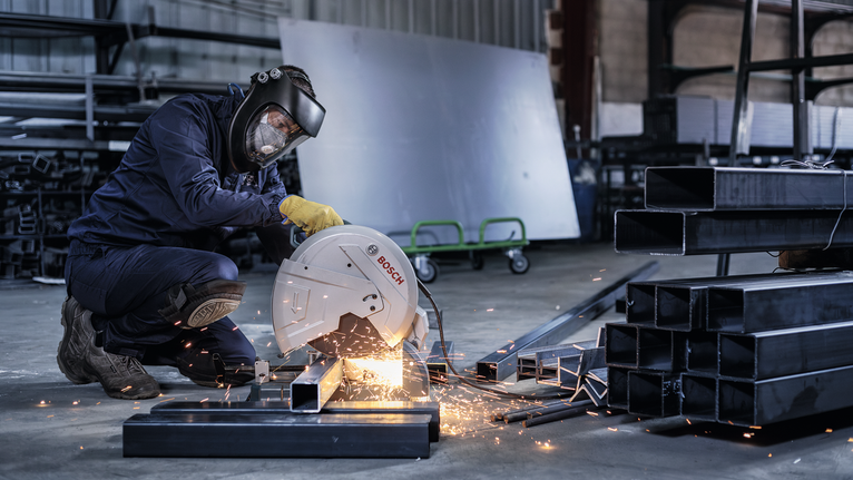 A worker wearing safety equipment cuts steel beams with a metal chop saw.