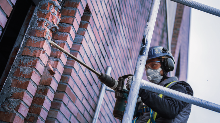 PRO SDS max-4C Pointed Chisel, 300 mm A person wearing safety equipment drills into a brick wall on scaffolding.