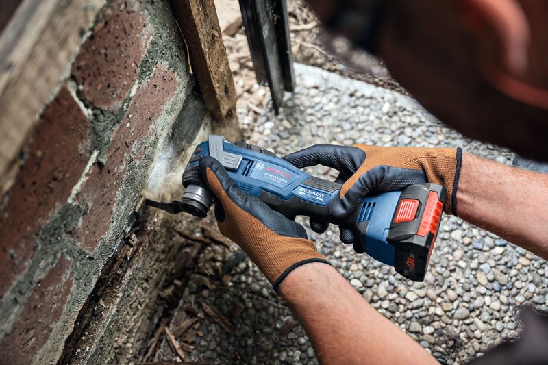 A person wearing safety equipment uses a cordless multi-cutter to cut into mortar near bricks.