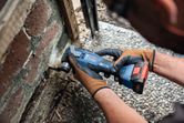 A person wearing safety equipment uses a cordless multi-cutter to cut into mortar near bricks.