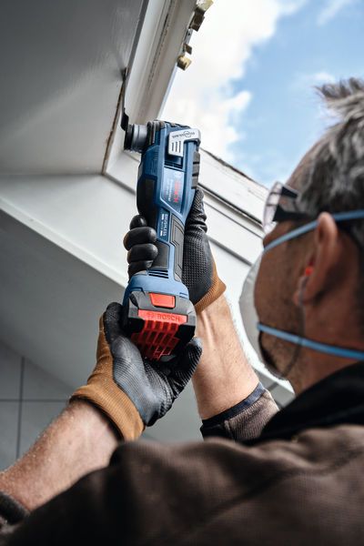 A person wearing safety equipment uses a cordless multi-cutter on a ceiling edge.
