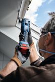 A person wearing safety equipment uses a cordless multi-cutter on a ceiling edge.