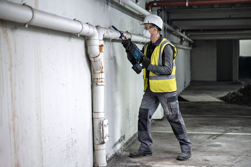 A person wearing safety equipment cuts a pipe on a wall with a reciprocating saw.