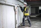 A person wearing safety equipment cuts a pipe on a wall with a reciprocating saw.