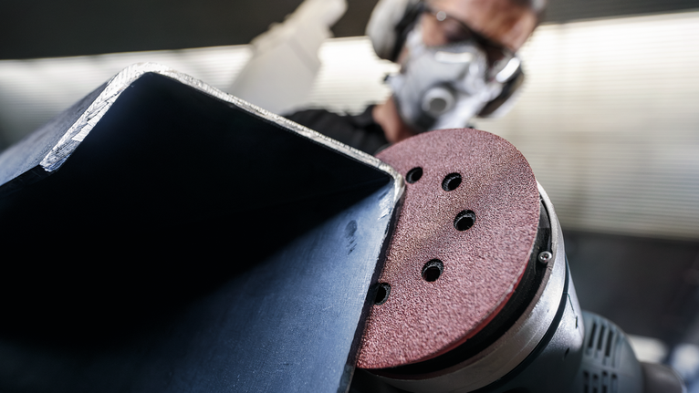 Person wearing safety equipment sands a metal beam with a power sander.