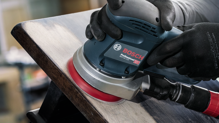 Person wearing safety equipment sands a wooden surface with an electric sander.