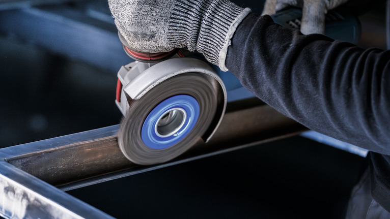 PRO Stainless Steel heavy X-Lock Wire Wheel, 115 x 0.5 mm Person wearing safety equipment grinds a metal beam with an angle grinder.