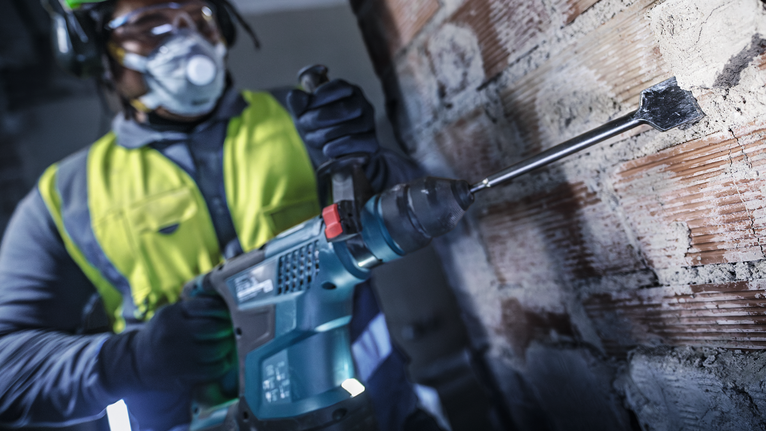 A person wearing safety equipment uses a rotary hammer to chisel a brick wall.